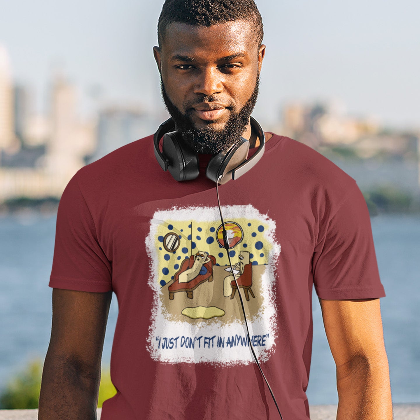 Man wearing a burgundy t-shirt with a graphic design and text, standing outdoors with a blurred cityscape in the background. A funny cartoon t-shirt for all you 'square pegs in round holes' out there. You are not alone. This unique design is ideal for anyone who thinks outside the box. Made from 100% organic ring-spun cotton, this witty cartoon t-shirt is unisex, high-quality, super comfy, and best of all, eco-friendly.