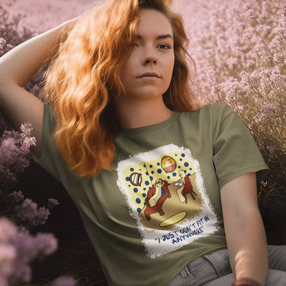 A woman with long wavy red hair wearing an khaki green graphic t-shirt in a field of pink flowers. The t-shirt graphic features a cartoon illustration of a person sitting on a lounge chair in a theorists room , with a white distressed border and the text 'I JUST DON'T FIT IN ANYWHERE' below.