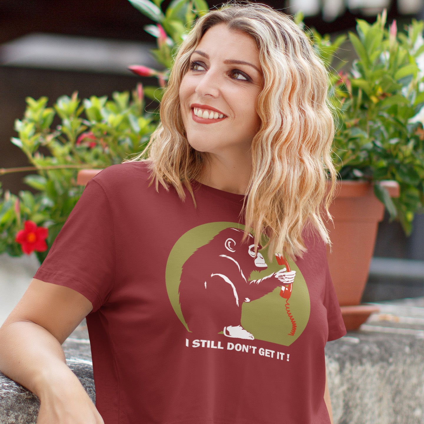 Woman wearing a burgundy t-shirt with a graphic design and text, sitting outdoors with plants in the background. This anti smart phone abuse t-shirt design highlights the human obsession with smart phones and how strange it must look to an ape. An outsiders view of a world glued to mobile phones. Made from 100% organic ring-spun cotton, this anti mobile phone abuse t-shirt is unisex, high-quality, super comfy, and best of all, eco-friendly.