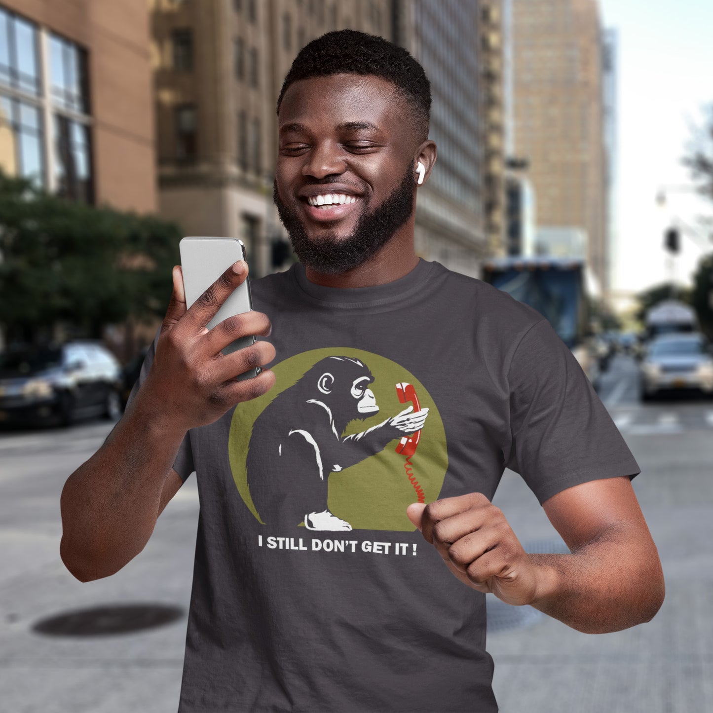 Man wearing a t-shirt with a graphic design of a monkey holding a phone and text 'I STILL DON'T GET IT!' on a city street. This satirical Banksy style stencil art t-shirt design highlights the human obsession with smart phones and how strange it must look to an ape. An outsiders view of a world glued to mobile phones. Made from 100% organic ring-spun cotton, this street art social awareness t-shirt is unisex, high-quality, super comfy, and best of all, eco-friendly.