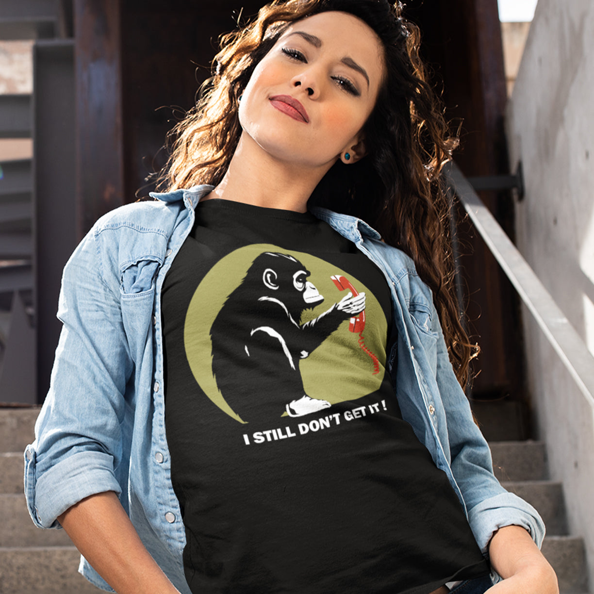 Woman wearing a black t-shirt with a graphic design and text, sitting on steps. This satirical Banksy style stencil art t-shirt design highlights the human obsession with smart phones and how strange it must look to an ape. An outsiders view of a world glued to mobile phones. Made from 100% organic ring-spun cotton, this street art social awareness t-shirt is unisex, high-quality, super comfy, and best of all, eco-friendly.