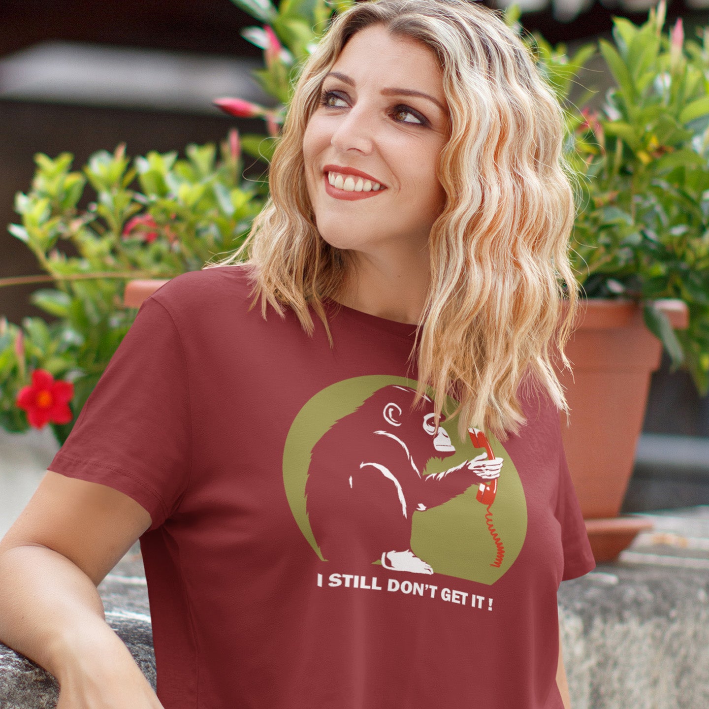 Woman wearing a burgundy t-shirt with a graphic design and text, standing outdoors with plants in the background. This satirical Banksy style stencil art t-shirt design highlights the human obsession with smart phones and how strange it must look to an ape. An outsiders view of a world glued to mobile phones. Made from 100% organic ring-spun cotton, this street art social awareness t-shirt is unisex, high-quality, super comfy, and best of all, eco-friendly.