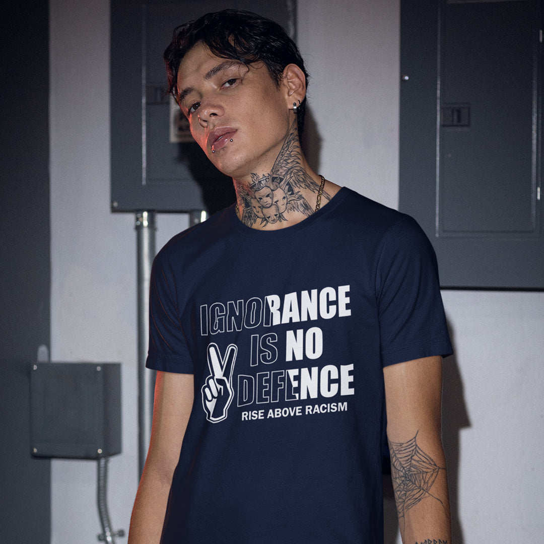 A man wearing a French navy blue graphic t-shirt with the text 'IGNORANCE IS NO DEFENCE RISE ABOVE RACISM' and a peace sign hand graphic, standing in front of grey electrical cabinets.