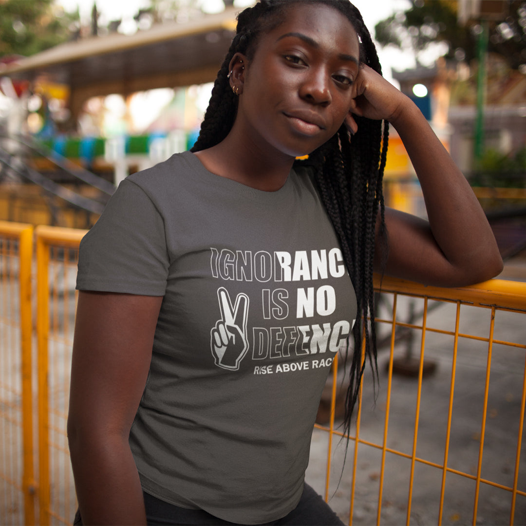 A woman wearing a dark gray graphic t-shirt with the text 'IGNORANCE IS NO DEFENSE' and a peace sign graphic, posing outdoors in front of a yellow fence.