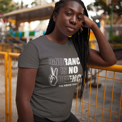 A woman wearing a dark gray graphic t-shirt with the text 'IGNORANCE IS NO DEFENSE' and a peace sign graphic, posing outdoors in front of a yellow fence.