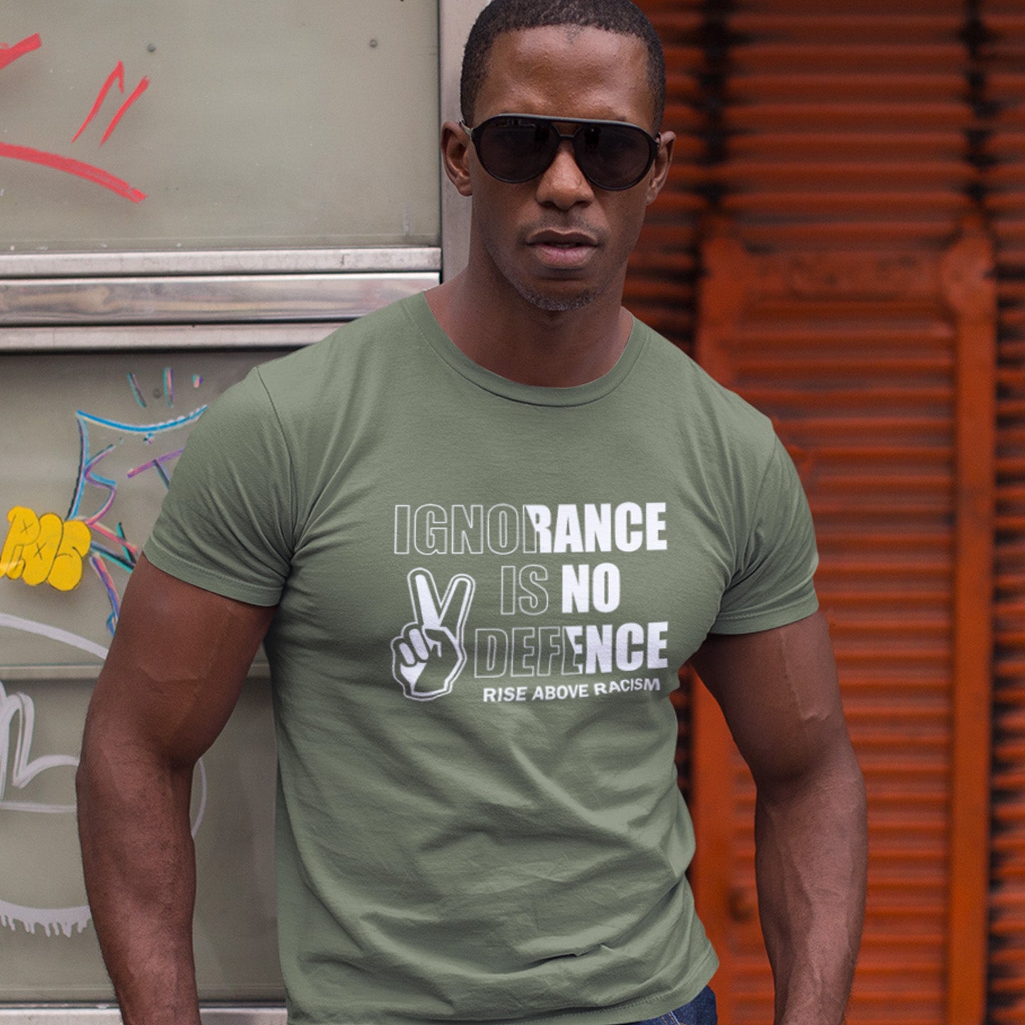 Man wearing a khaki t-shirt with a message in front of a graffiti-covered wall. An anti-racist t-shirt to remind people that ignorance is no defence against prejudice. This unique racial equality t-shirt design promotes understanding and brotherhood to achieve peace in the world. Made from 100% organic ring-spun cotton, this anti racism t-shirt is unisex, high-quality, super comfy, and best of all, eco-friendly.