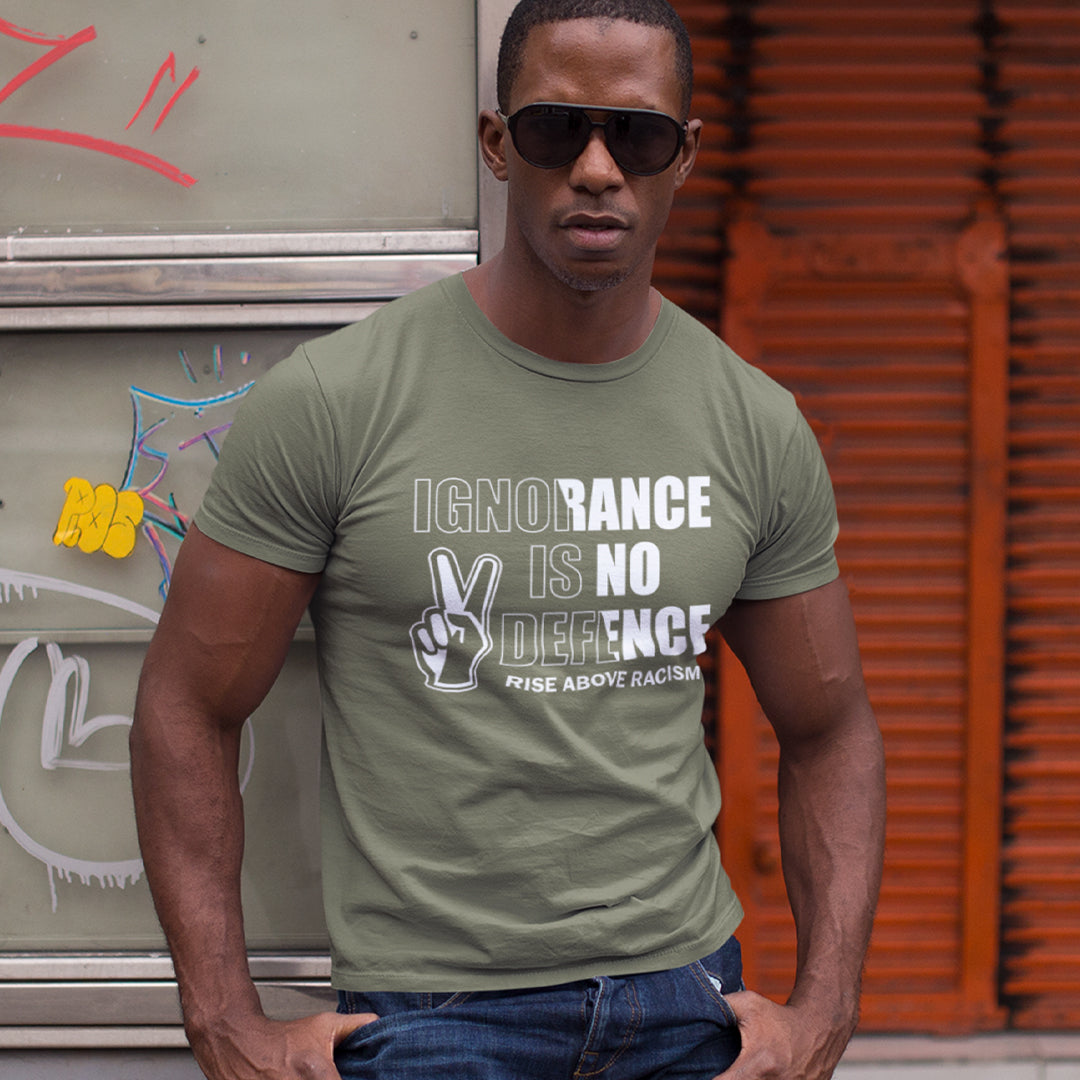 A man wearing an olive green graphic t-shirt with the text 'IGNORANCE IS NO DEFENSE' and a peace sign graphic, paired with dark sunglasses and blue jeans, standing in front of a graffiti-covered wall and an orange corrugated metal shutter.
