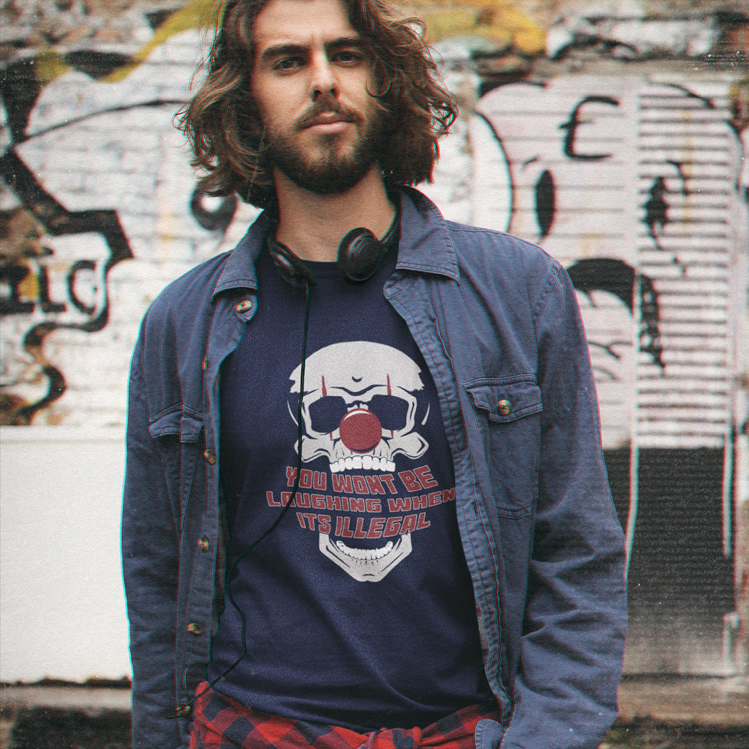 A man with long brown hair and a beard stands in front of a graffiti-covered wall, wearing a French navy blue graphic t-shirt with a skull and football design, a blue denim jacket, and a red plaid shirt.