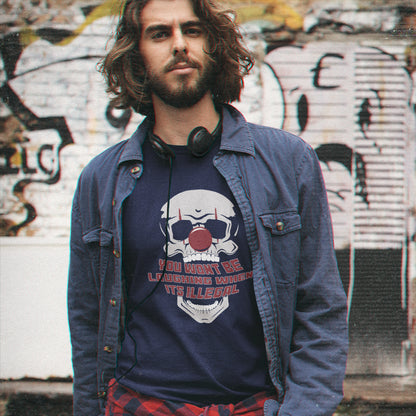 A man with long brown hair and a beard stands in front of a graffiti-covered wall, wearing a French navy blue graphic t-shirt with a skull and football design, a blue denim jacket, and a red plaid shirt.