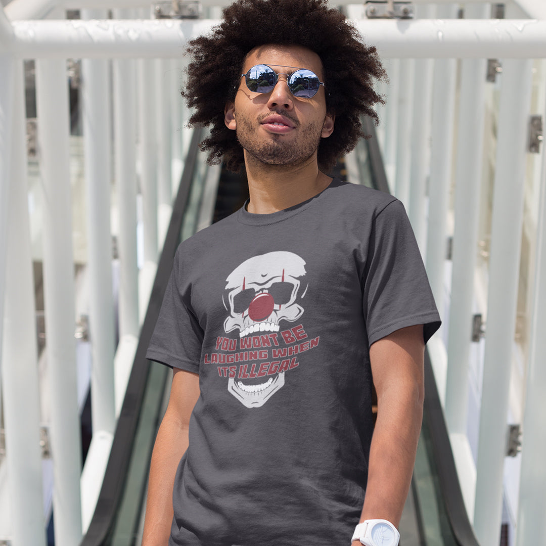 Man standing near a bridge, wearing an anthracite grey t-shirt featuring a graphic of a skull wearing a red clown nose, with the text 'YOU WON'T BE LAUGHING WHEN IT'S ILLEGAL' in red and white outlined lettering below.