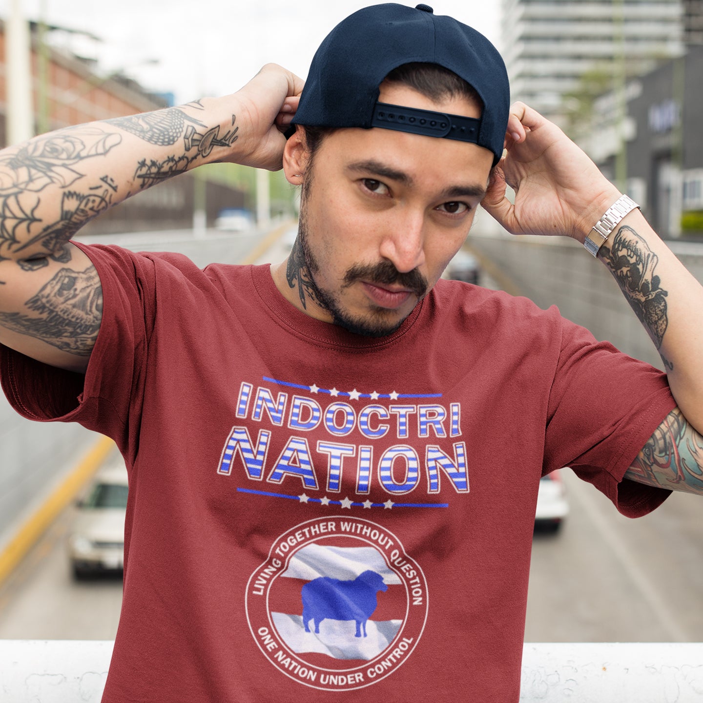 Man wearing a red t-shirt with 'Indoctrination' text and graphic, outdoors. A political t-shirt with satirical look at nationalism, an insight into a very dark future for any nation under a controlling government. Display your social awareness with this unique design. Made from 100% organic ring-spun cotton, this political t-shirt is unisex, high-quality, super comfy, and best of all, eco-friendly.