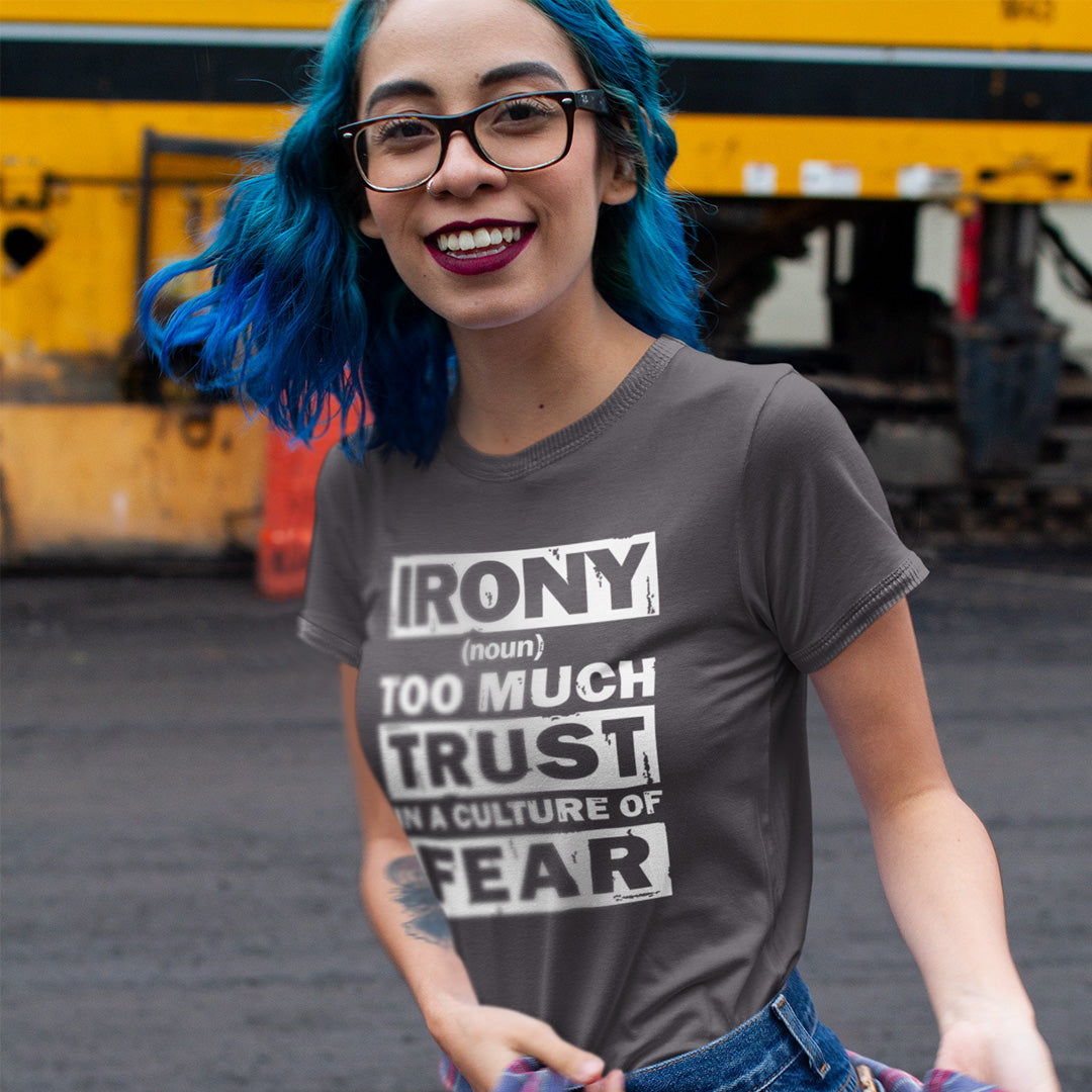 A woman with bright blue hair and black glasses wearing a dark gray graphic t-shirt that reads 'IRONY (noun) TOO MUCH TRUST IN A CULTURE OF FEAR'. She is standing outdoors in front of a yellow train.