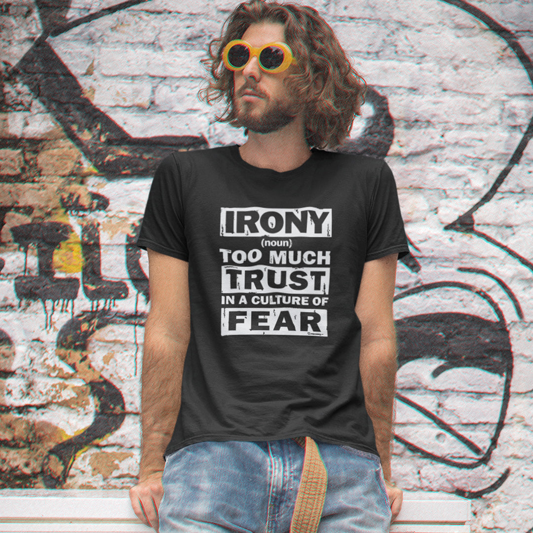 A man with long curly hair and yellow sunglasses wearing a black graphic t-shirt that reads 'IRONY (noun) TOO MUCH TRUST IN A CULTURE OF FEAR'. He is standing in front of a graffiti-covered brick wall.