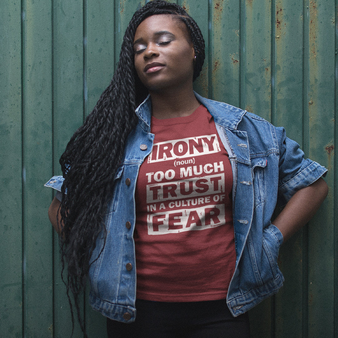 A woman with long black braids stands against a green corrugated metal wall, wearing a red graphic t-shirt under an open blue denim jacket. The t-shirt features a white block text graphic that reads 'IRONY (noun) TOO MUCH TRUST IN A CULTURE OF FEAR'.