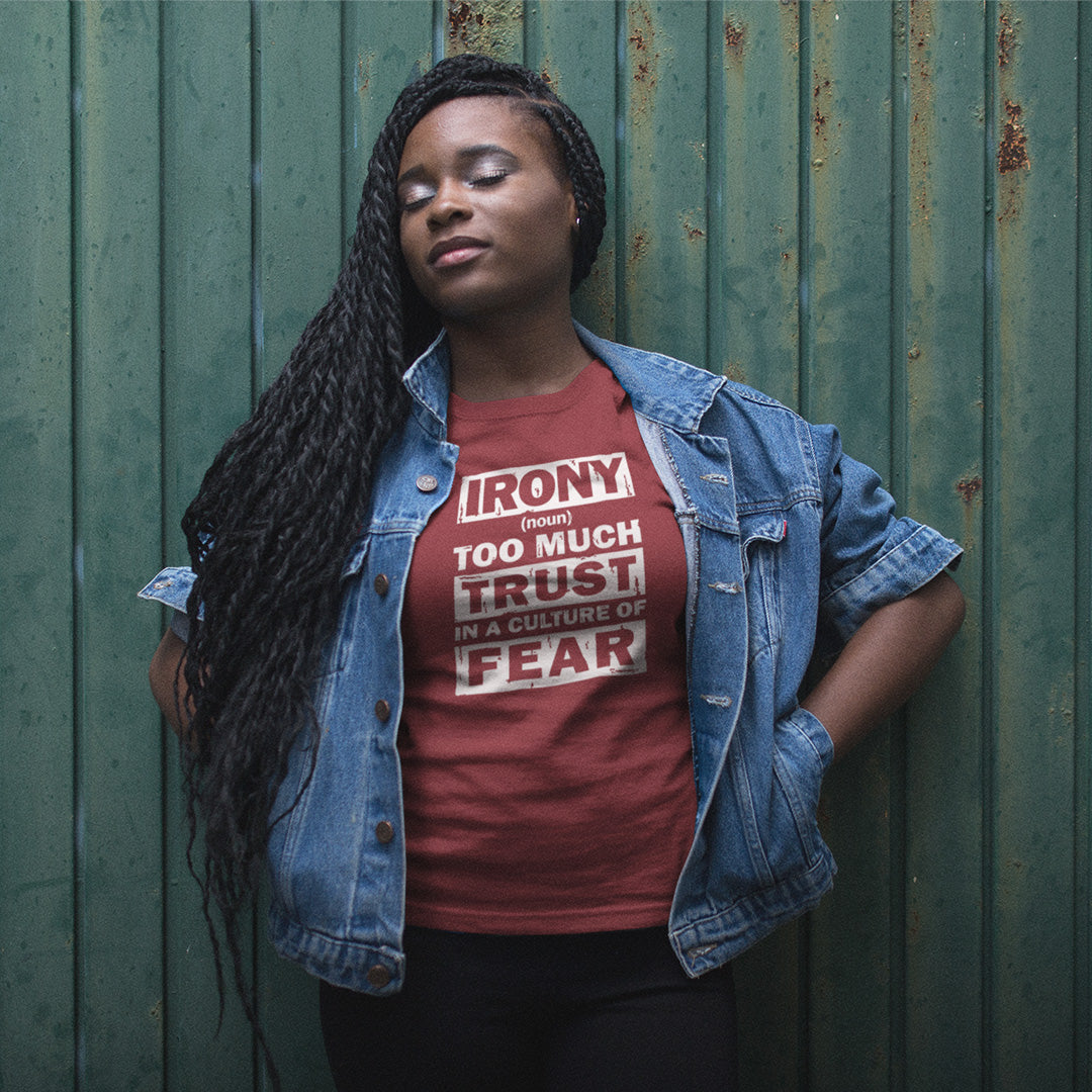 A woman with long black braids stands against a weathered green metal wall, wearing a red graphic t-shirt and a light blue denim jacket. The t-shirt features a white graphic that reads 'IRONY from TOO MUCH TRUST IN A CULTURE OF FEAR'.