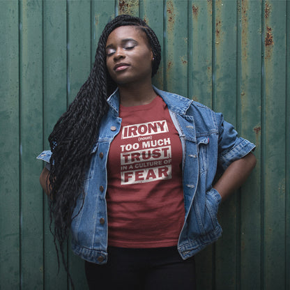 A woman with long black braids stands against a weathered green metal wall, wearing a red graphic t-shirt and a light blue denim jacket. The t-shirt features a white graphic that reads 'IRONY from TOO MUCH TRUST IN A CULTURE OF FEAR'.