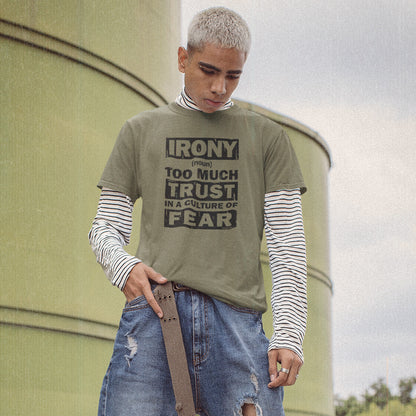 A man with platinum blonde hair wearing an olive green graphic t-shirt with a black text print, layered over a black and white striped long-sleeve shirt, paired with distressed blue jeans and a brown belt, standing in front of a large olive-green cylindrical structure.