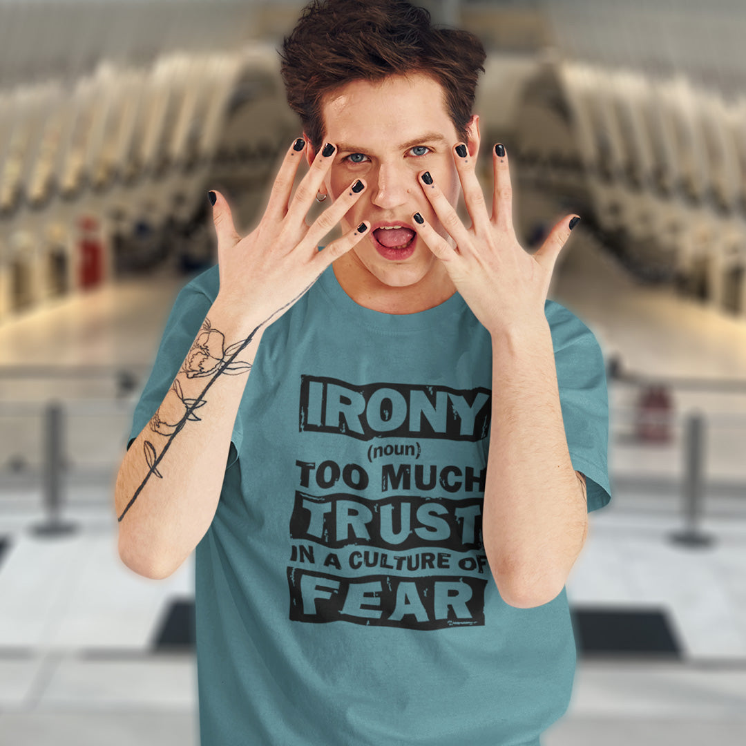 A man wearing a teal graphic t-shirt with the text 'IRONY (noun) TOO MUCH TRUST IN A CULTURE OF FEAR' in black. He is posing with his hands near his face, showing black fingernails, in a blurred indoor setting.