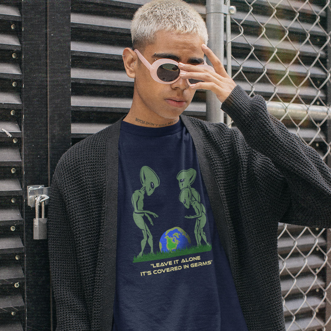A person wearing a French navy graphic t-shirt with a green alien design and text, styled with a black knit cardigan and light pink sunglasses, standing in front of a black metal gate and chain-link fence.