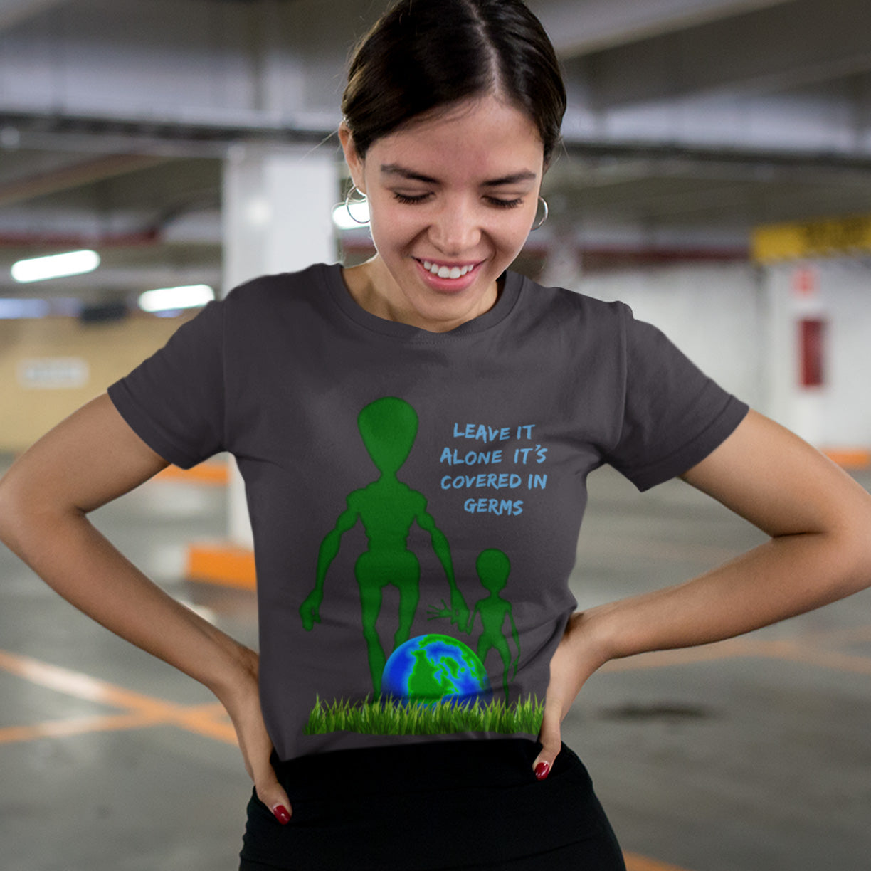 Woman wearing a t-shirt with an alien graphic and text in a parking garage. A funny alien t-shirt design with a subtle overpopulation message. In the grand scheme of things, our planet and universe could just be the school project of a child from a superior race. Made from 100% organic ring-spun cotton, this funny sci fi t shirt is unisex, high-quality, super comfy, and best of all, eco-friendly.