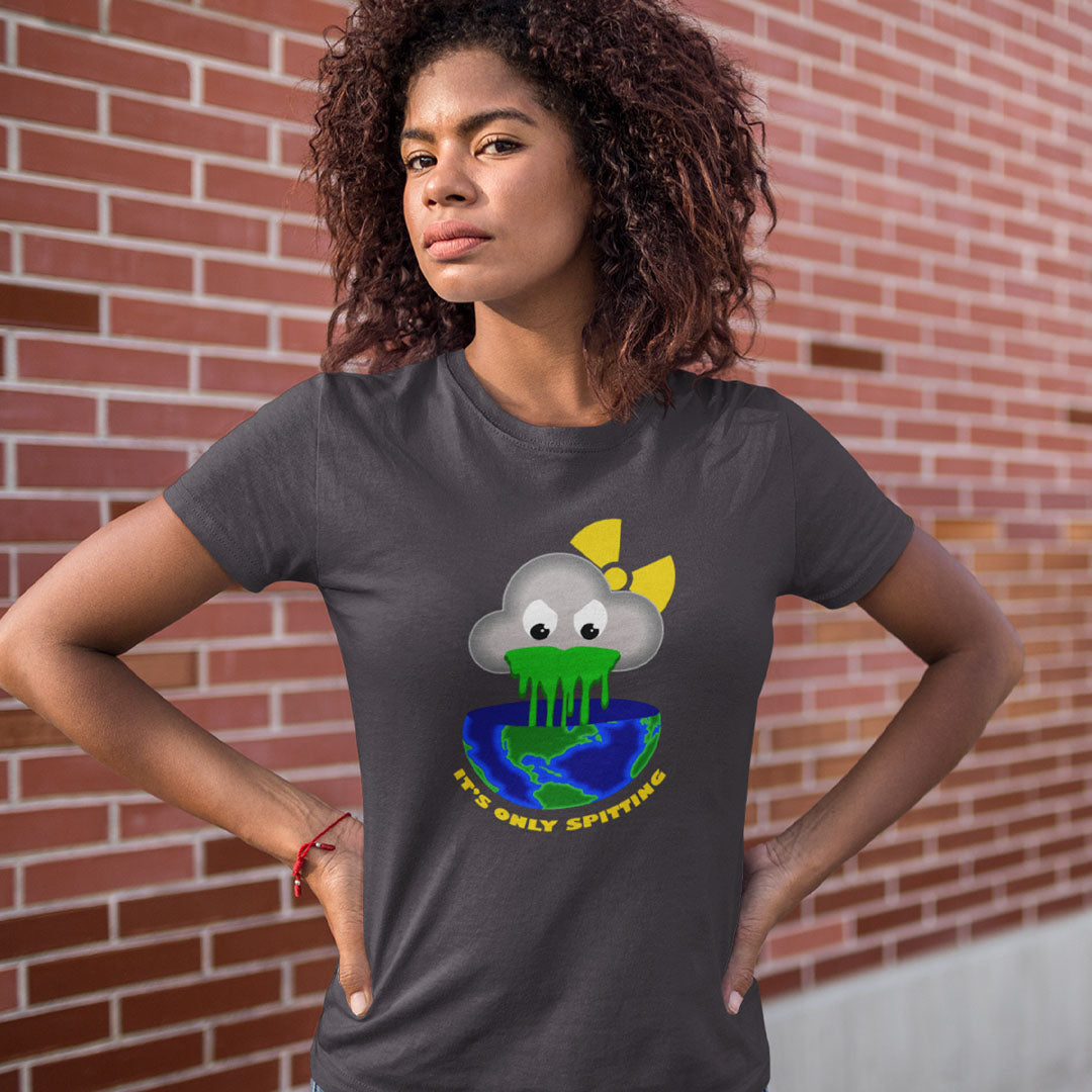 A woman with curly brown hair stands with hands on hips in front of a red brick wall, wearing a dark gray t-shirt with a cartoon graphic of a cloud planet spitting green acid onto a blue and green Earth, with the text 'IT'S ONLY SPITTING'.