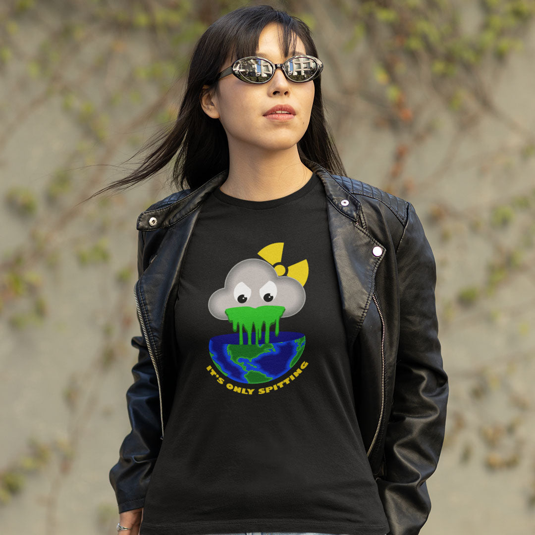 A woman wearing a black graphic t-shirt and a black leather jacket, standing outdoors against a blurred background of beige stone walls and green foliage. The t-shirt features a cartoon cloud character with a yellow bow, green slime, and a blue/green globe, with the text 'IT'S ONLY SPITTING'.