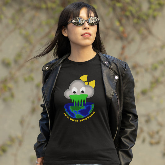 A woman wearing a black graphic t-shirt and a black leather jacket, standing outdoors against a blurred background of beige stone walls and green foliage. The t-shirt features a cartoon cloud character with a yellow bow, green slime, and a blue/green globe, with the text 'IT'S ONLY SPITTING'.