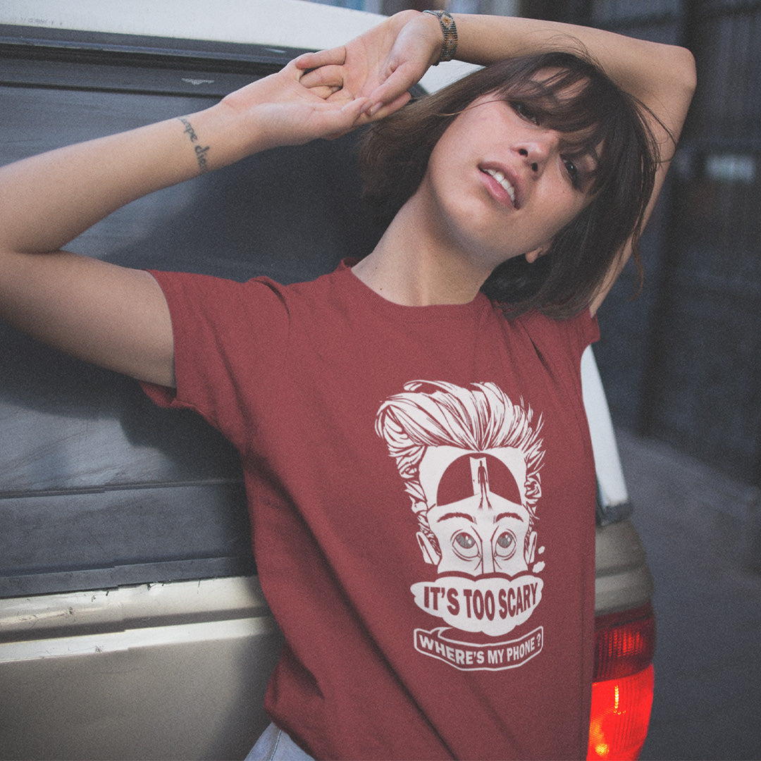 A woman with raised arms, standing at the back end of a car, wearing a burgundy short-sleeve crewneck t-shirt featuring a white graphic print of a cartoon face with wild hair and the text 'IT'S TOO SCARY WHERE'S MY PHONE?' on the front, displayed against a solid white background.