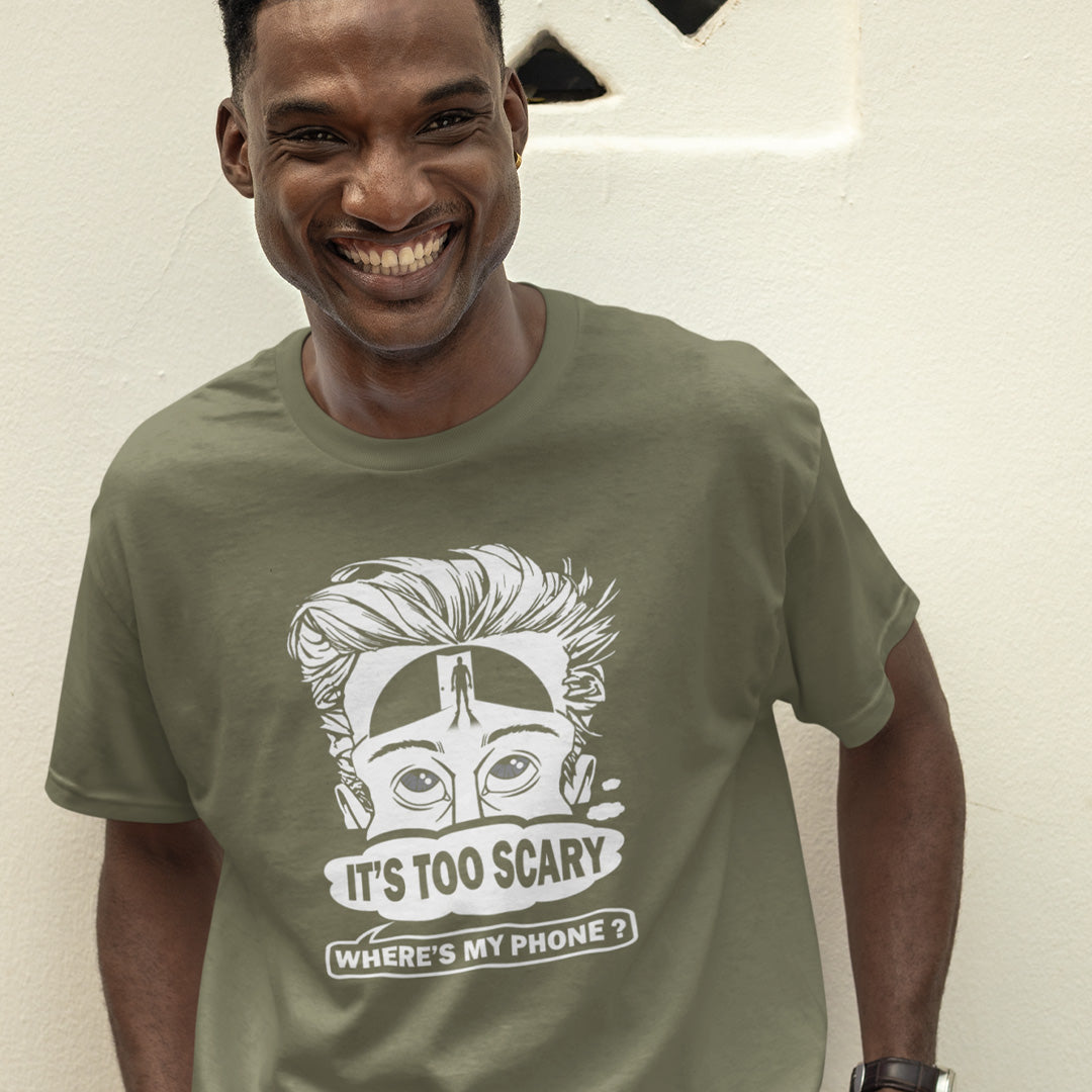 A laughing man standing outside, wearing a khaki green  short-sleeve crewneck t-shirt featuring a white graphic print of a cartoon face with wild hair and the text 'IT'S TOO SCARY WHERE'S MY PHONE?' on the front, displayed against a solid white background.