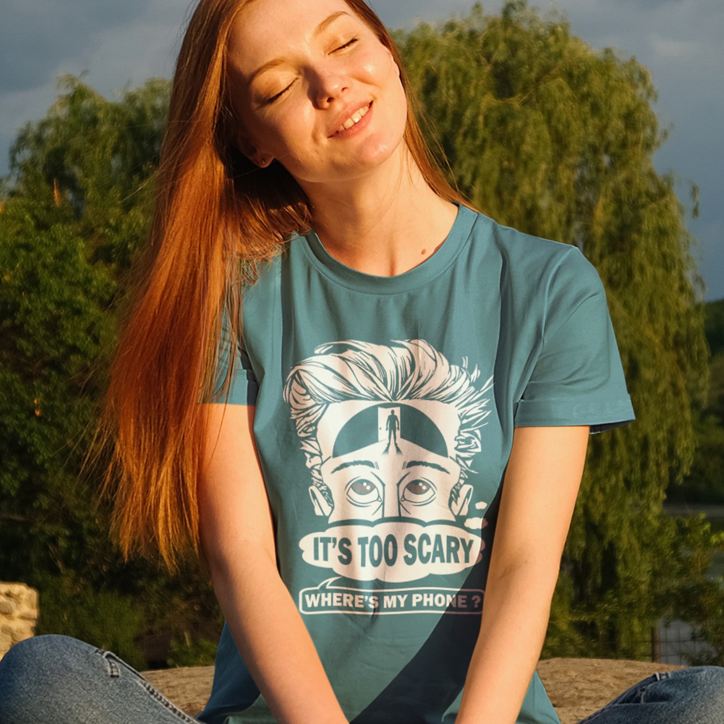Woman wearing a stargazer t-shirt with a graphic design and text, sitting outdoors. An anti-mobile phone abuse t-shirt highlighting the effects on the mind of smart phone over use in the place of thinking. A view of how mobile phones have taken over thinking and left us with dark unexplored corridors in our minds. Made from 100% organic ring-spun cotton, this social awareness t-shirt is unisex, high-quality, super comfy, and best of all, eco-friendly.