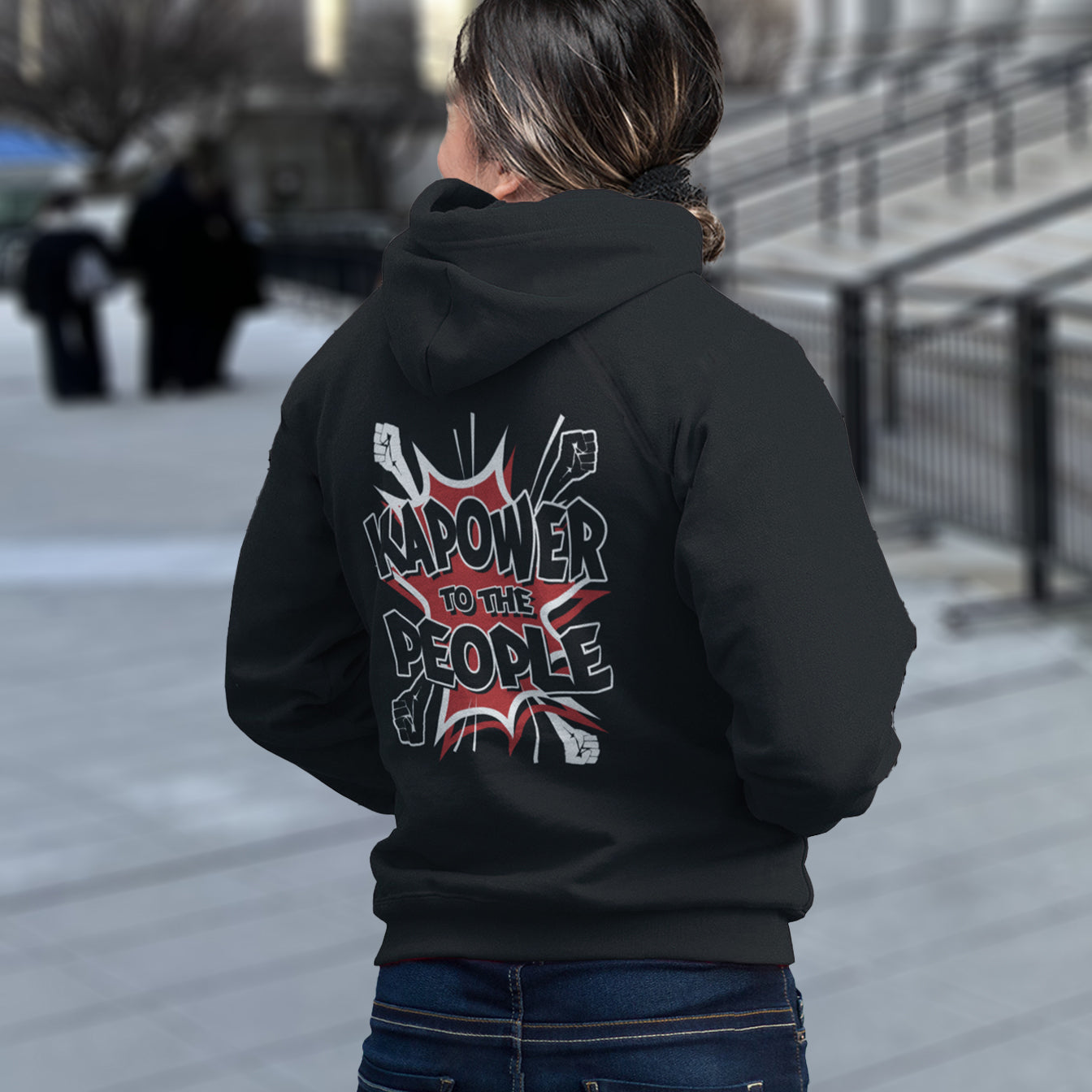 Person wearing a black hoodie with 'Kapower to the People' design on the back, standing outdoors. An anti-government hoodie design that packs a punch with this humorous play on the slogan 'Power to the people', with a comic book twist. Support true democracy with this witty political message. Comfortable and practical. It's extra soft and has a convenient front pouch pocket. Made from organic cotton and recycled polyester, this social awareness hoodie is a great eco-friendly choice.