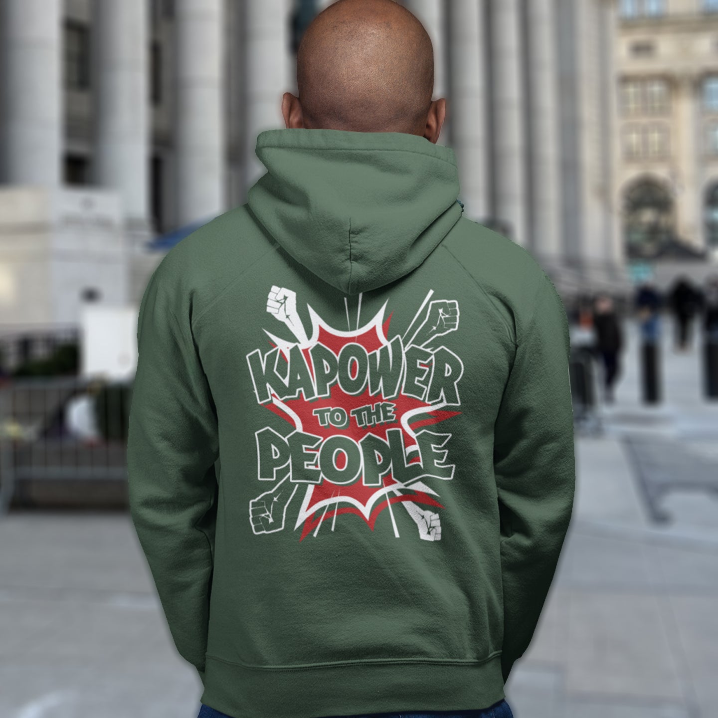 Person wearing a bottle green hoodie with 'Kapower to the People' text on the back, standing in an urban setting. An anti-government hoodie design that packs a punch with this humorous play on the slogan 'Power to the people', with a comic book twist. Support true democracy with this witty political message. Comfortable and practical. It's extra soft and has a convenient front pouch pocket. Made from organic cotton and recycled polyester, this social awareness hoodie is a great eco-friendly choice.