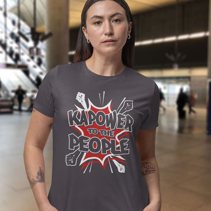 Person wearing a t-shirt with 'Kapower to the People' design in a blurred indoor setting. This anti government t shirt packs a punch with this humorous play on the slogan 'Power to the people', with a comic book twist. Support true democracy with this funny political message. Made from 100% organic ring-spun cotton, this unisex non conformist t shirt is a total must-have. It's high-quality, super comfy, and best of all, eco-friendly.