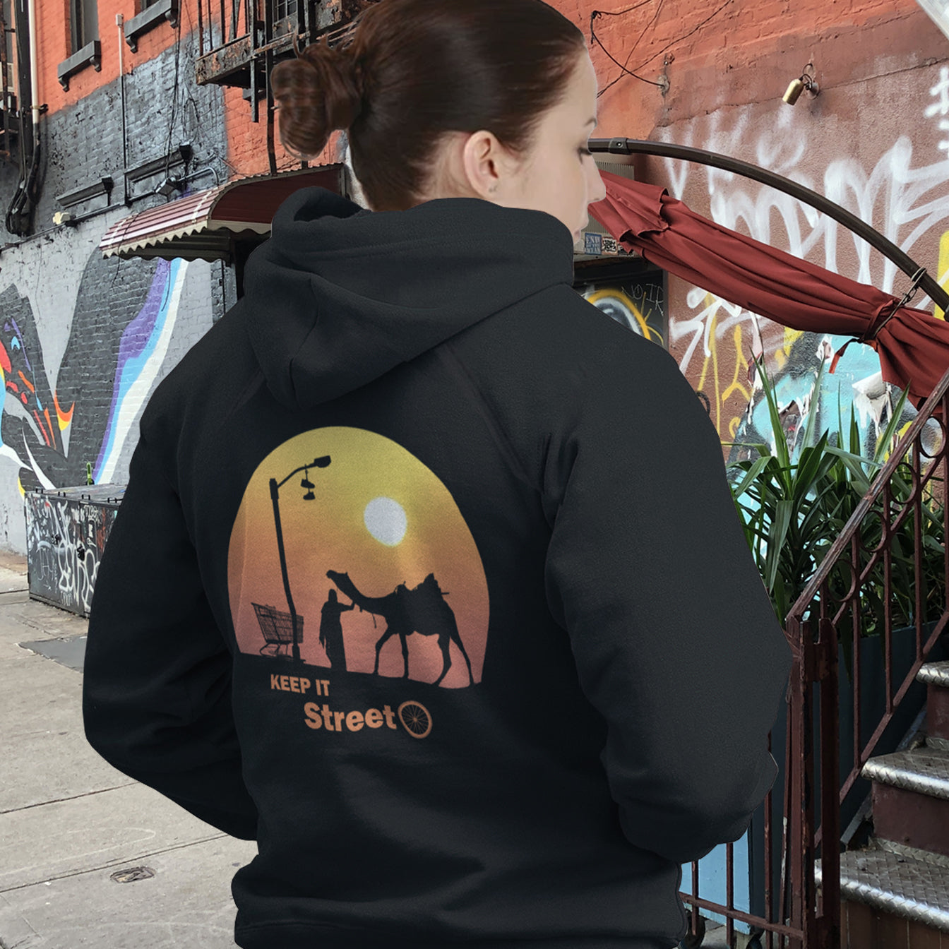 Person wearing a black hoodie with a graphic design on the back, standing in an urban setting with graffiti. This street art inspired hoodie design represents inner city street life as a culture of it's own.  Wherever you are in the world, may the street be with you. Comfortable and practical. It's extra soft and has a convenient front pouch pocket. Made from organic cotton and recycled polyester, this street art hoodie is a great eco-friendly choice.