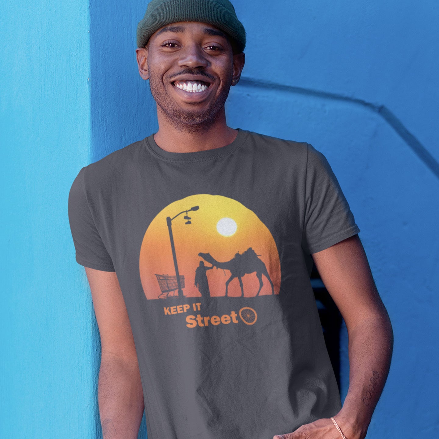 Man wearing a t-shirt with a graphic design of a sunset, camel, and people, against a blue background. This street art t shirt stencil represents inner city street life as a culture of it's own. There is a ground level reality mixed with trustworthy honestly at street level in towns and cities across the planet. Wherever you are in the world, may the street be with you. Made from 100% organic ring-spun cotton, this urban art t shirt is unisex, high-quality, super comfy, and best of all, eco-friendly.