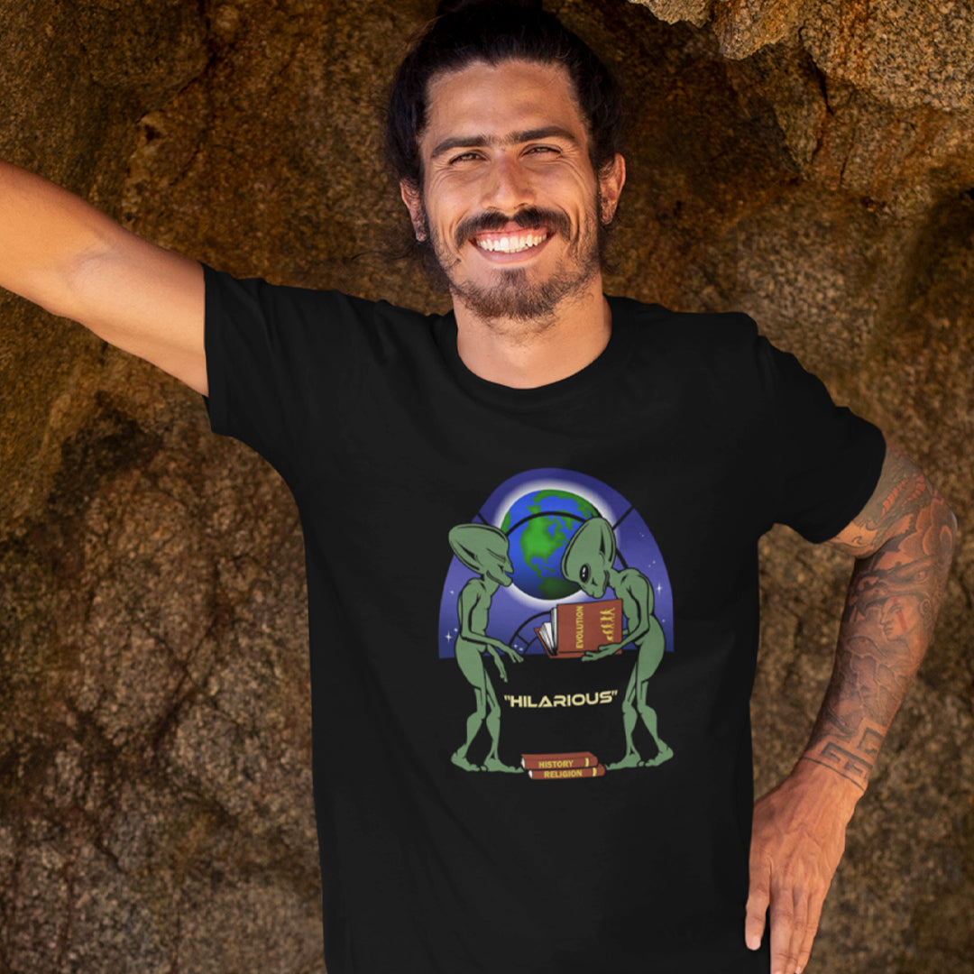 Man wearing a black t-shirt with a graphic design of an alien holding a book, standing against a stone wall. This funny alien t shirt suggest we are being lied to about our history and the aliens know the truth. A design for alien loving conspiracy theorists. Have the aliens had more influence in our lives than we are lead to believe ? Made from 100% organic ring-spun cotton, these cool sci fi t shirts are unisex, high-quality, super comfy, and best of all, eco-friendly.