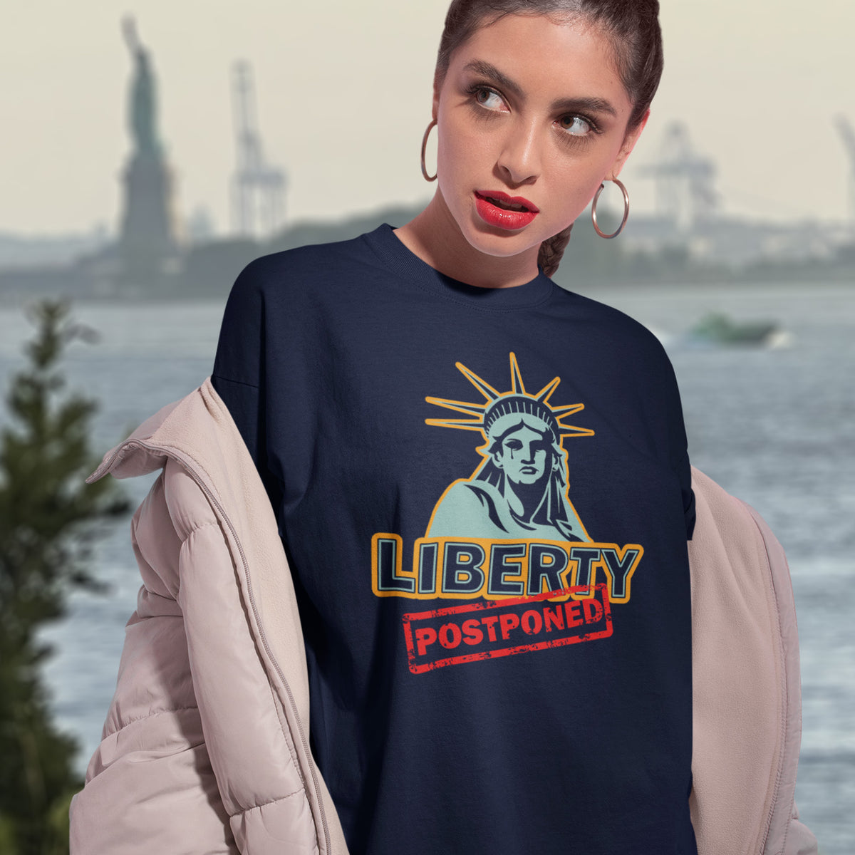 A woman with red lipstick standing in front of a view of the Statue of Liberty, wearing a French nay blue short sleeved crew neck t-shirt with a crying Statue of Liberty design with the text "LIBERTY POSTPONED" underneath.