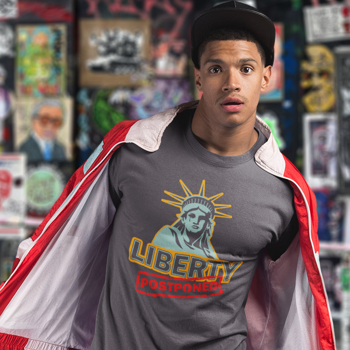 A surprised looking man, standing in front of a poster filled wall, wearing a black cap and an anthracite grey crew neck t-shirt with a crying Statue of Liberty design with the text "LIBERTY POSTPONED" underneath.
