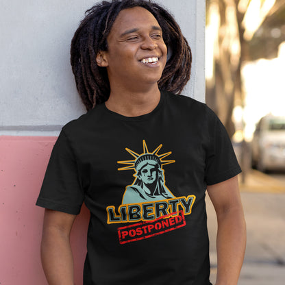 A smiling man with dreadlocks, leaning against a wall, wearing a black short sleeved crew neck t-shirt with a crying Statue of Liberty design with the text "LIBERTY POSTPONED" underneath.
