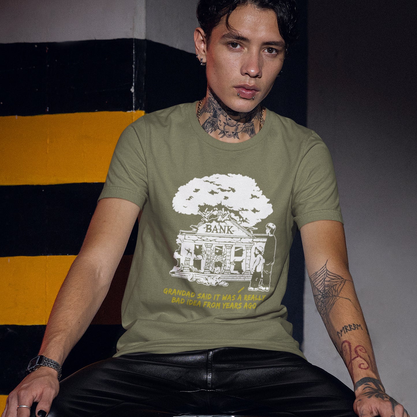 Person wearing a green t-shirt with a graphic design sitting against a striped wall. A Banksy style stencil art t-shirt with an anti-capitalism message. A thought provoking future where money and banking are a dark memory and our children have something more natural to look forward to. Made from 100% organic ring-spun cotton, this utopian graffiti stencil t-shirt is unisex, high-quality, super comfy, and best of all, eco-friendly.