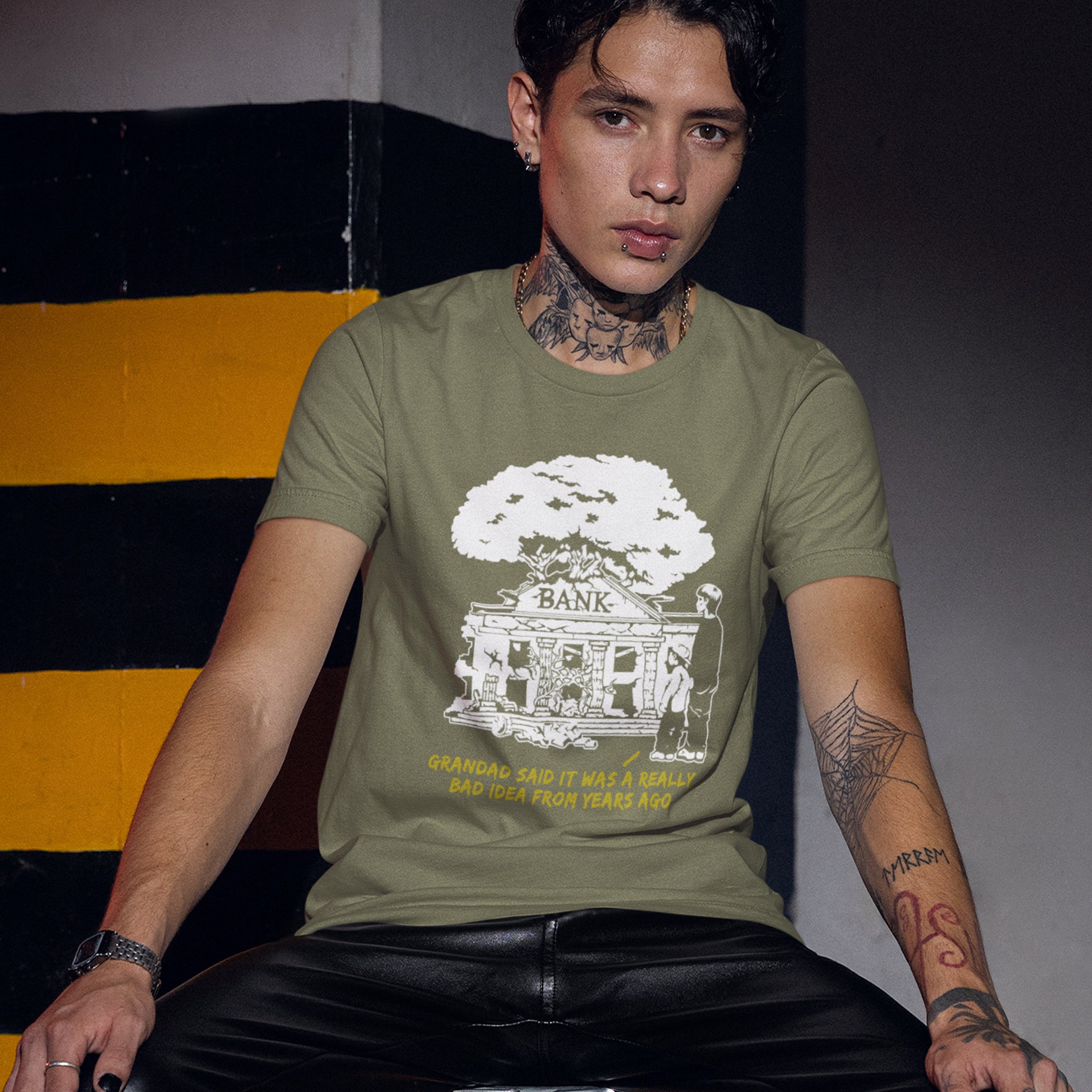 Person wearing a green t-shirt with a graphic design sitting against a striped wall. A Banksy style stencil art t-shirt with an anti-capitalism message. A thought provoking future where money and banking are a dark memory and our children have something more natural to look forward to. Made from 100% organic ring-spun cotton, this utopian graffiti stencil t-shirt is unisex, high-quality, super comfy, and best of all, eco-friendly.