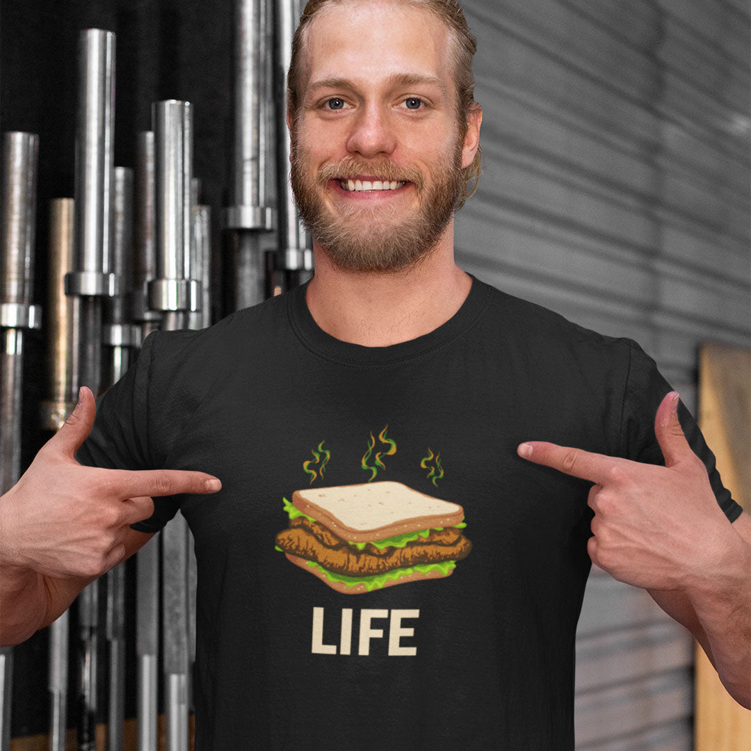 A smiling man with a beard points at a black graphic t-shirt featuring a cartoon sandwich with green steam lines and the word 'LIFE' printed below. The background shows a gym setting with weight bars on the left and a gray corrugated wall on the right.