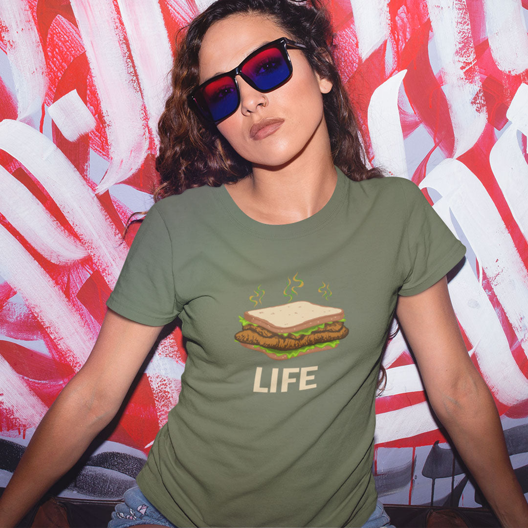 A woman wearing an olive green graphic t-shirt with a burger and the word 'LIFE' printed on the front, paired with red and blue 3D-effect sunglasses, posing in front of a red and white abstract background.