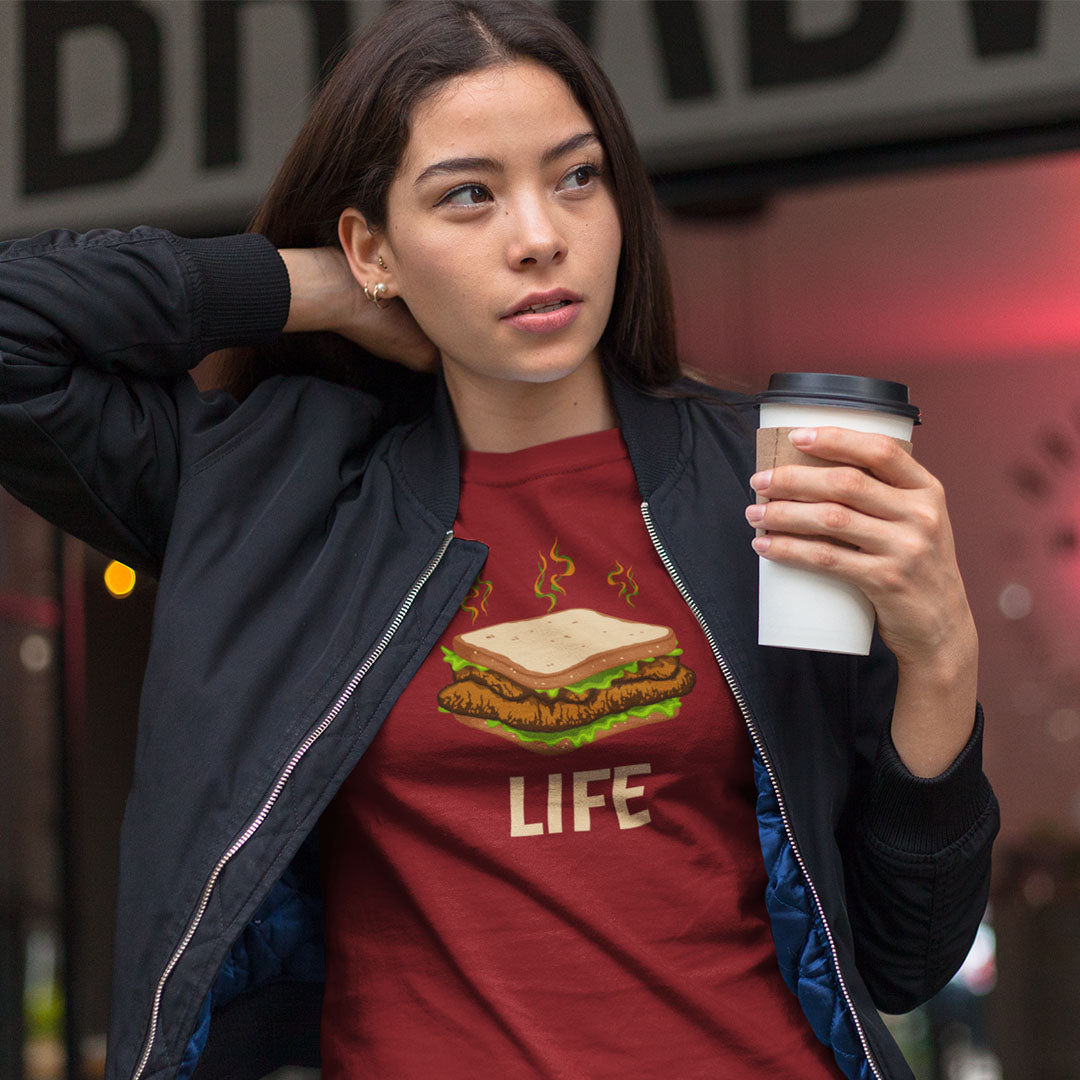 A woman wearing a red graphic t-shirt with a burger and the word 'LIFE' printed on the front, styled with an open black bomber jacket and holding a white coffee cup.