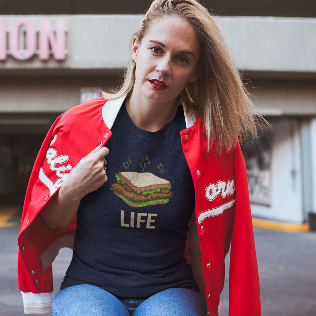 A woman wearing a navy graphic t-shirt with a burger and the word 'LIFE' printed on the front, layered under an open red varsity jacket with white lettering, posed in front of a blurred street background.