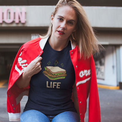 A woman wearing a navy graphic t-shirt with a burger and the word 'LIFE' printed on the front, layered under an open red varsity jacket with white lettering, posed in front of a blurred street background.