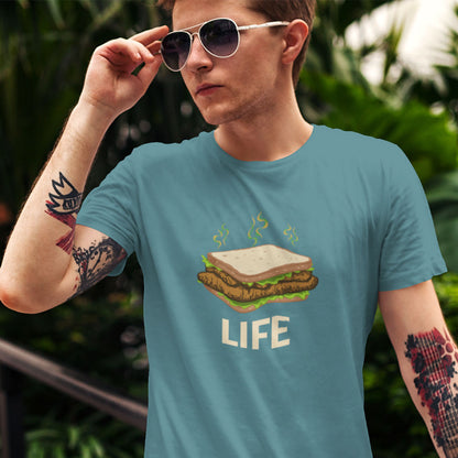 A man wearing a light blue graphic t-shirt with a burger and the word 'LIFE' printed on the front, set against a blurred green outdoor background.