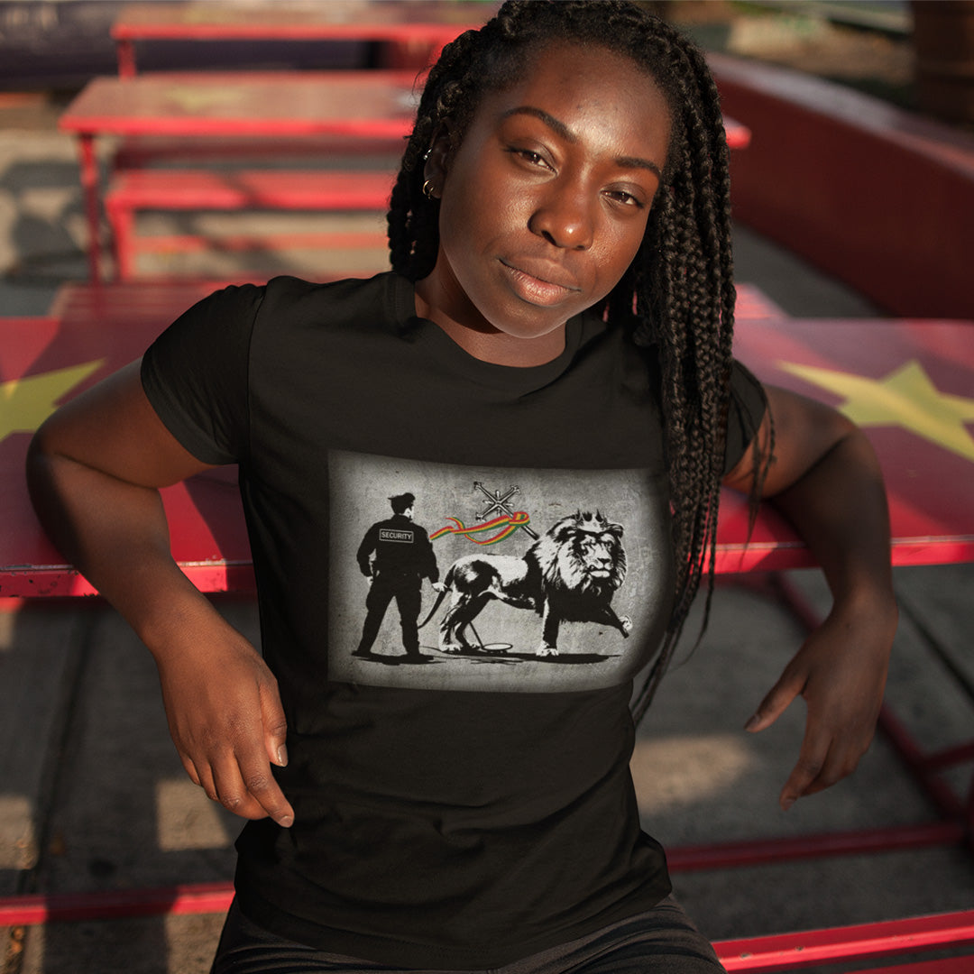 A woman with platted hair leaning back against a table, wearing a black short sleeved crew neck t-shirt with a graffiti stencil design of a security guard checking the underside of the Lion of Judah for explosives.