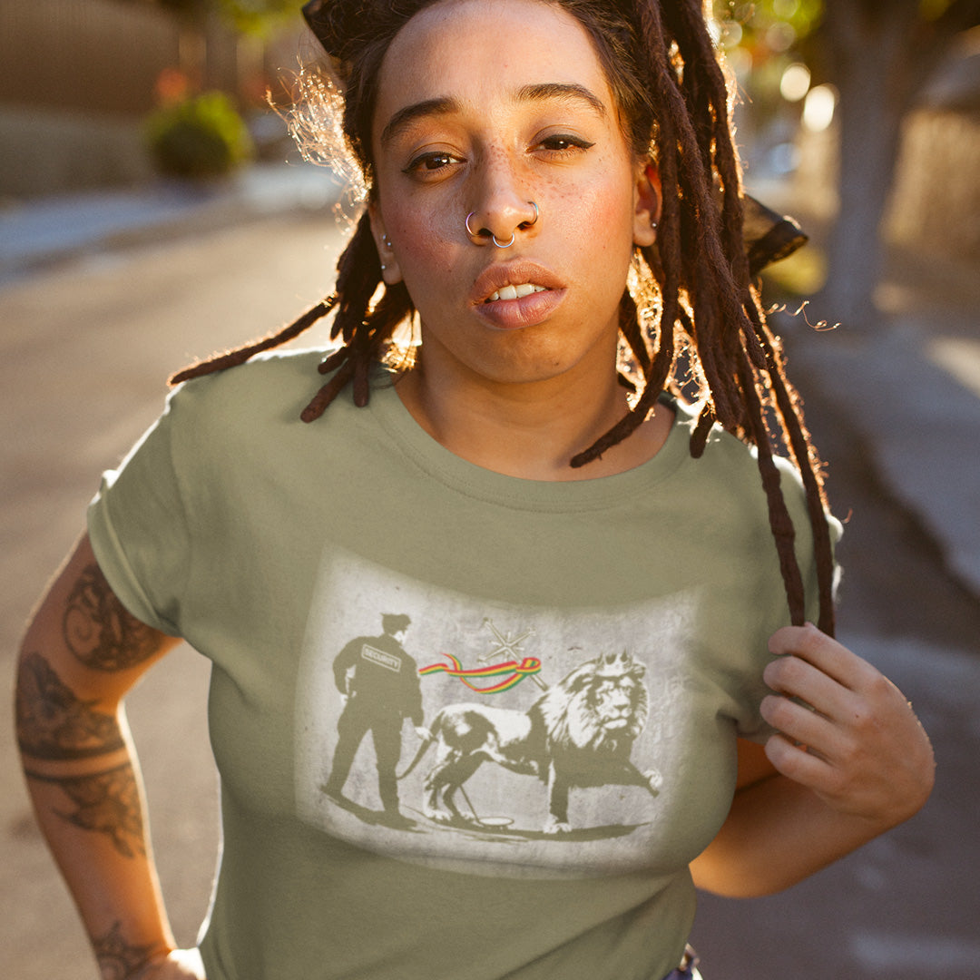 A woman with dreadlocks standing in the street, wearing a khaki short sleeved crew neck t-shirt with a graffiti stencil design of a security guard checking the underside of the Lion of Judah for explosives.