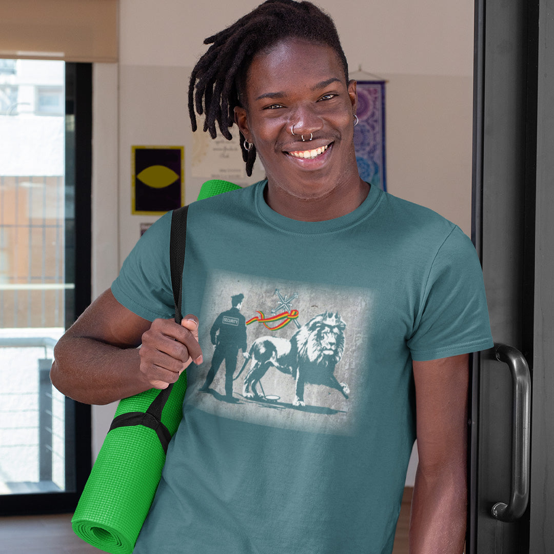 A man with dreadlocks and piercings holding a yoga mat and wearing a stargazer blue short sleeved crew neck t-shirt with a graffiti stencil design of a security guard checking the underside of the Lion of Judah for explosives.